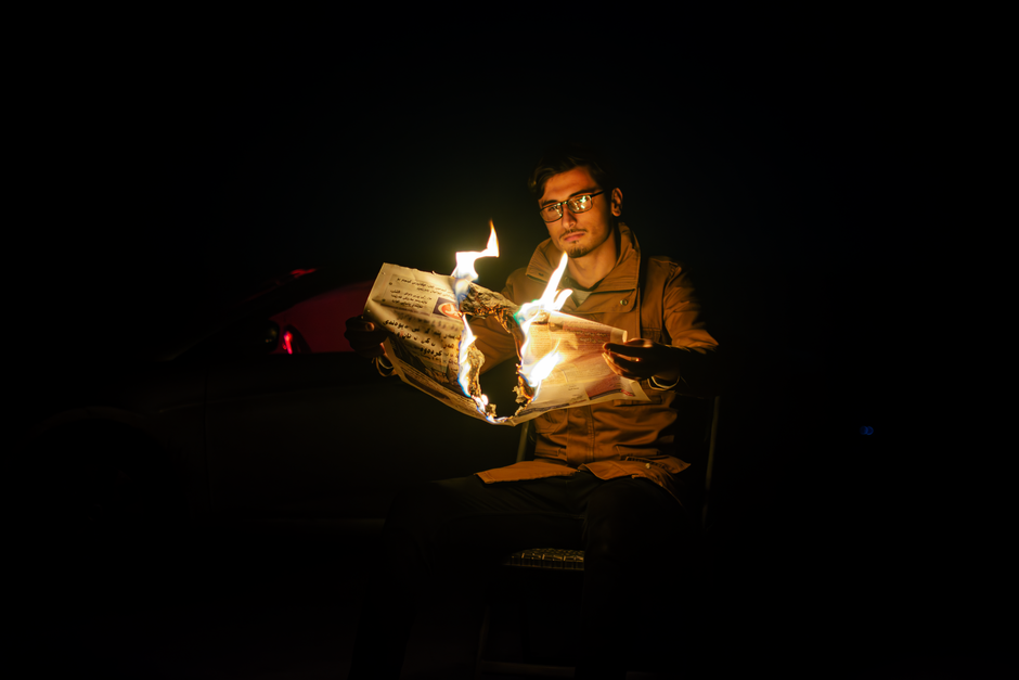 Moody image of a man sitting alone at night, burning a newspaper, creating a dramatic effect.