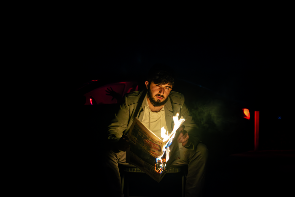 A man sitting outdoors at night reads a burning newspaper, creating a dramatic, moody scene.