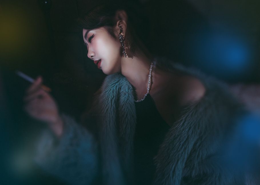 Stylish woman in fur coat and jewelry smoking in moody lighting.