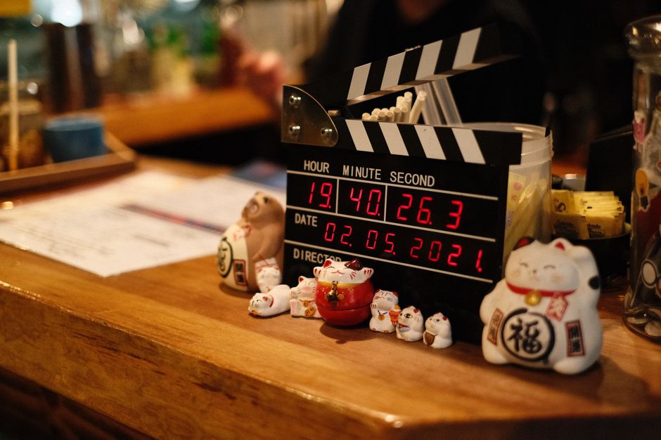 A close-up of Maneki Neko figurines on a wooden table beside a clapperboard and digital clock in a cozy setting.