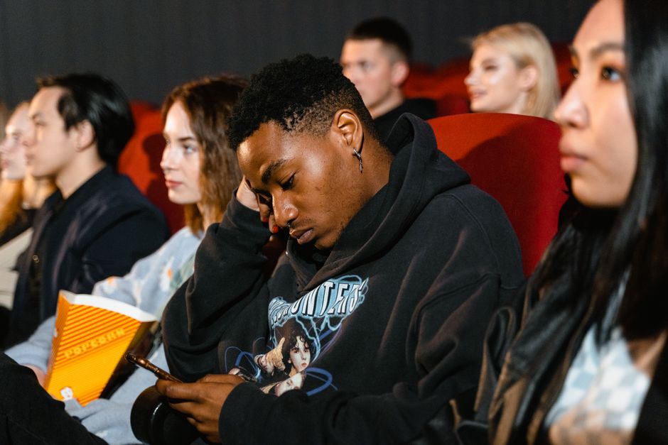 A diverse group of young adults in a cinema, focused on their smartphones and popcorn.