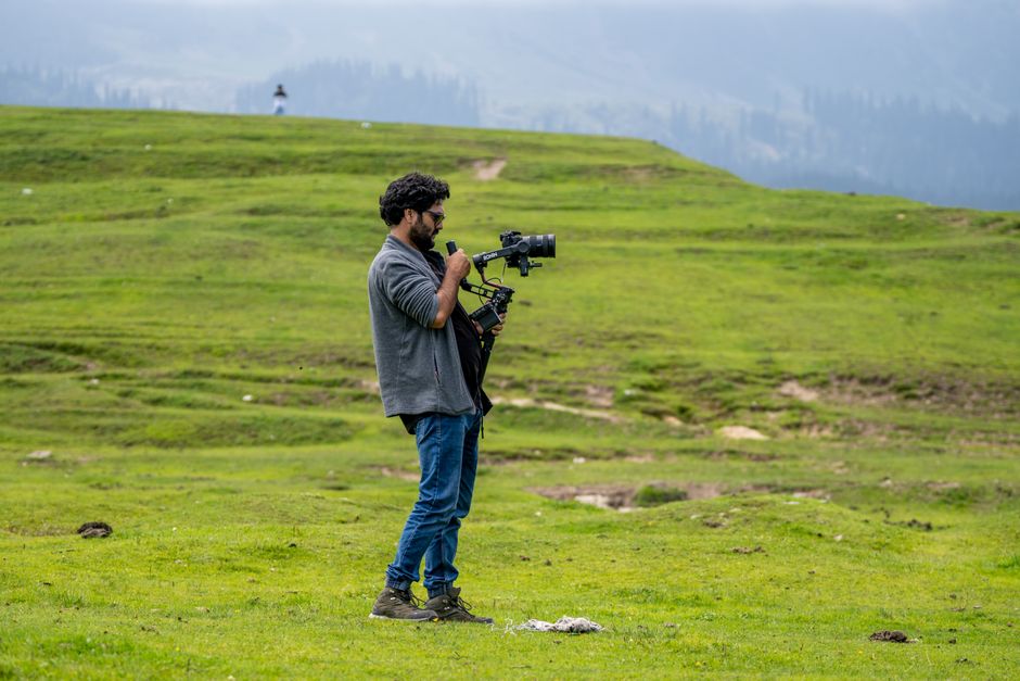 A videographer with a gimbal captures footage in a picturesque green meadow, showcasing outdoor filmmaking.