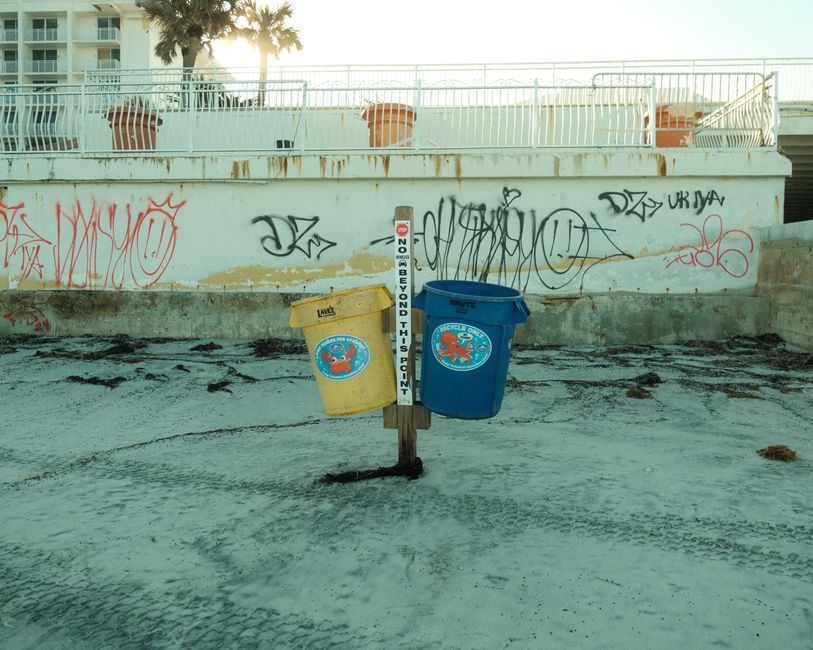 Graffiti-covered wall with blue and yellow recycling bins on a sandy beach.
