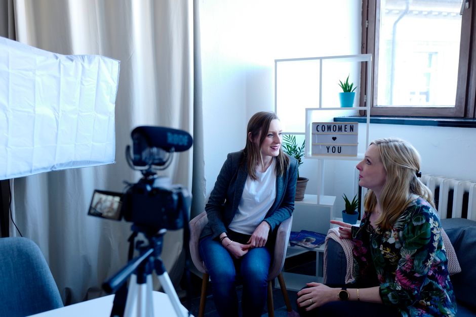 Two women discuss together in a modern workspace setting with camera equipment.