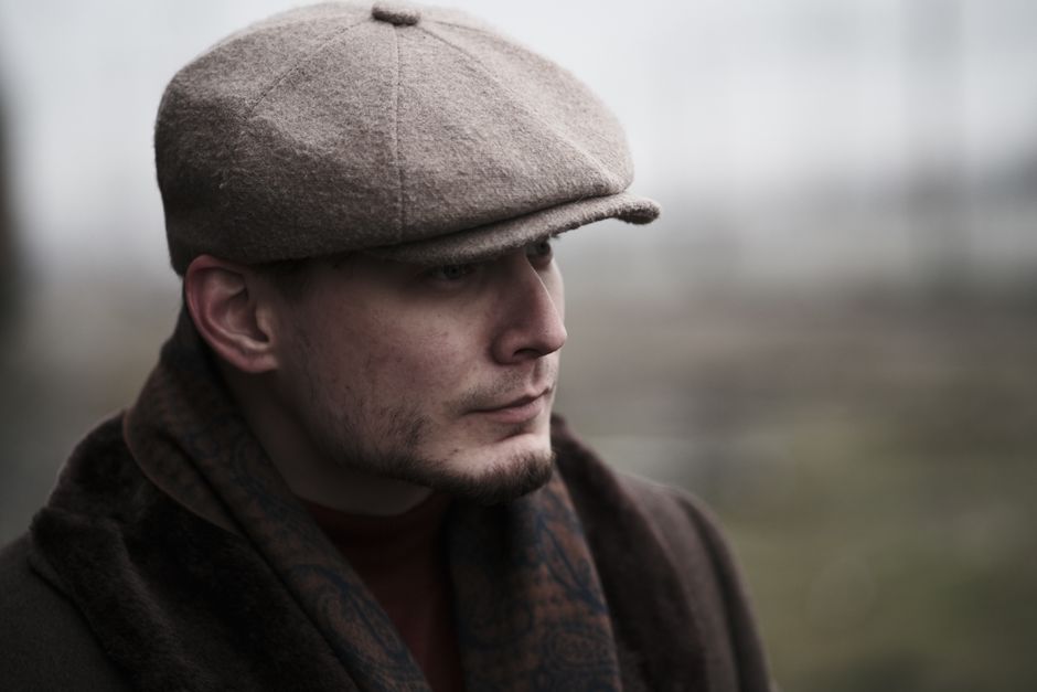Profile portrait of a man wearing a flat cap and coat, looking thoughtful outdoors.