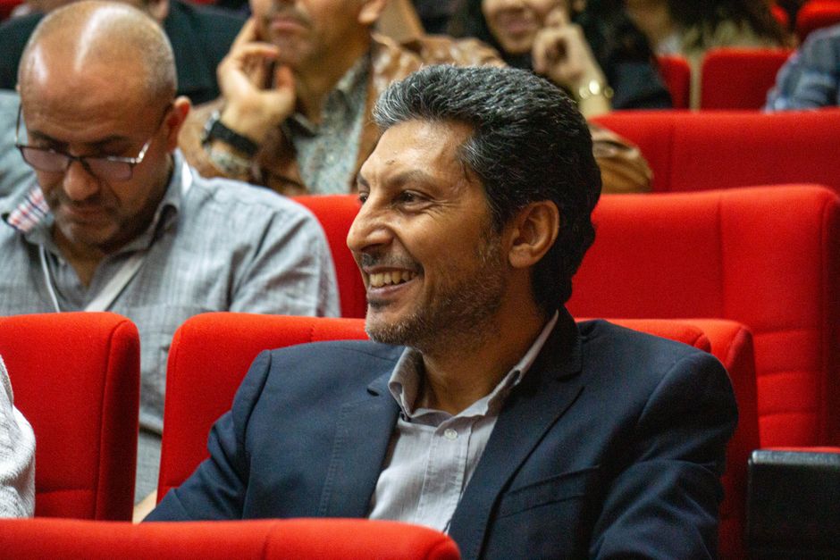 A man in a suit smiles while attending a conference, seated in a red auditorium.