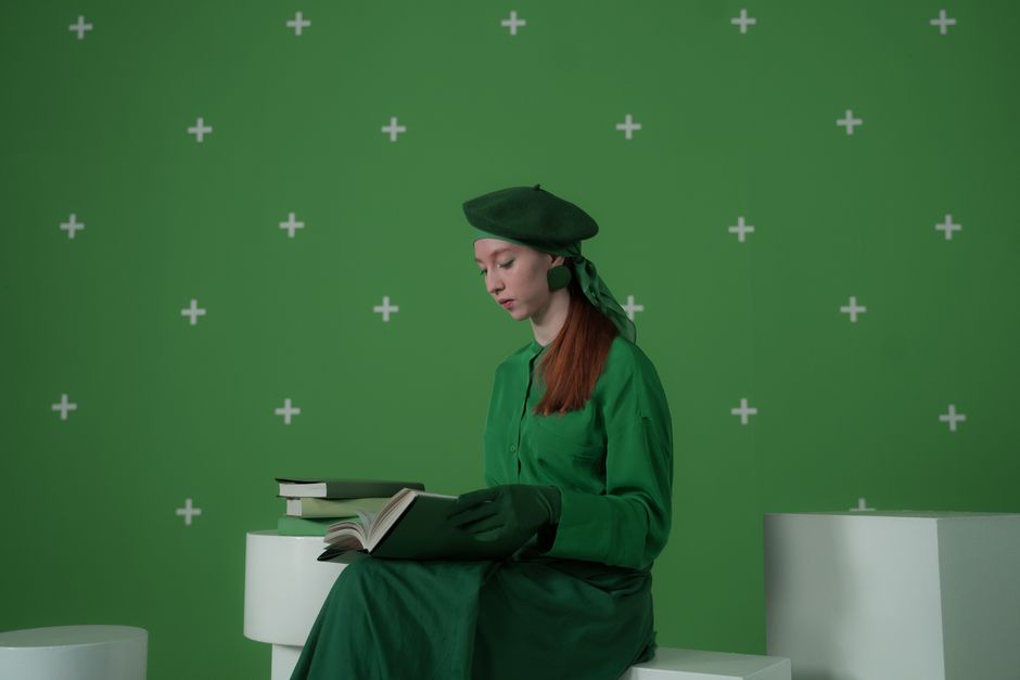 A woman in green reads a book in front of a green screen. Indoor setting.