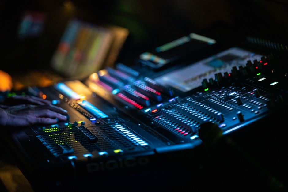 Close-up of hands operating a digital audio mixing console with colorful LEDs and blurred background.