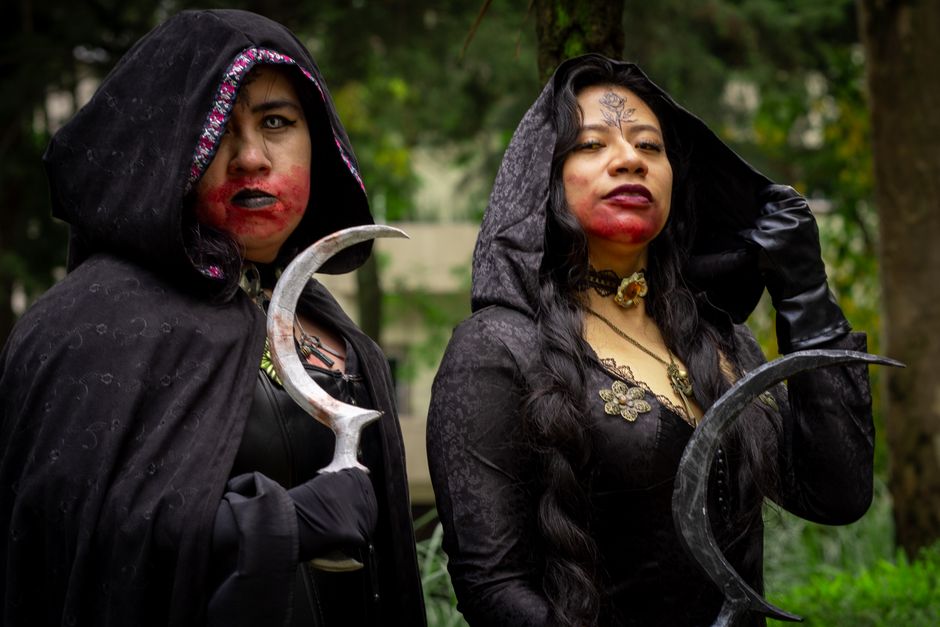 Two women in dark cosplay with dramatic makeup holding scythes outdoors.
