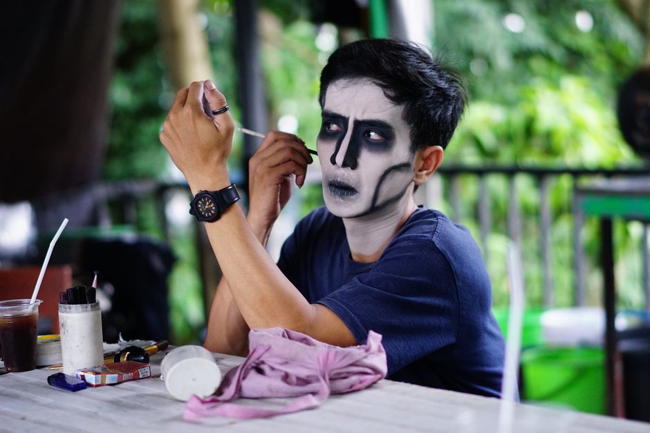 A man applying dark, gothic face paint outdoors, surrounded by makeup tools and greenery.