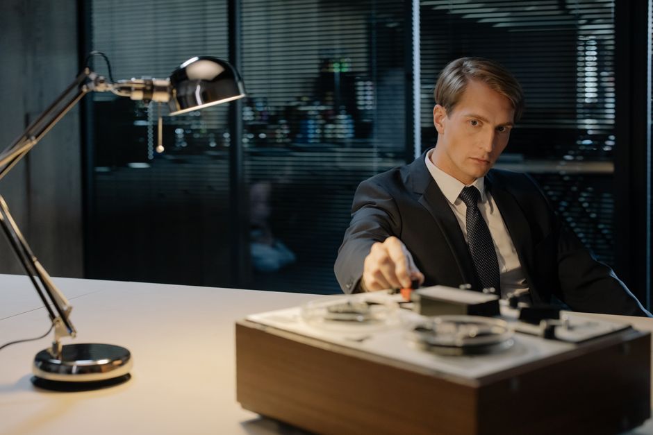 A detective in a suit uses a tape recorder under a desk lamp, evoking vintage investigation themes.