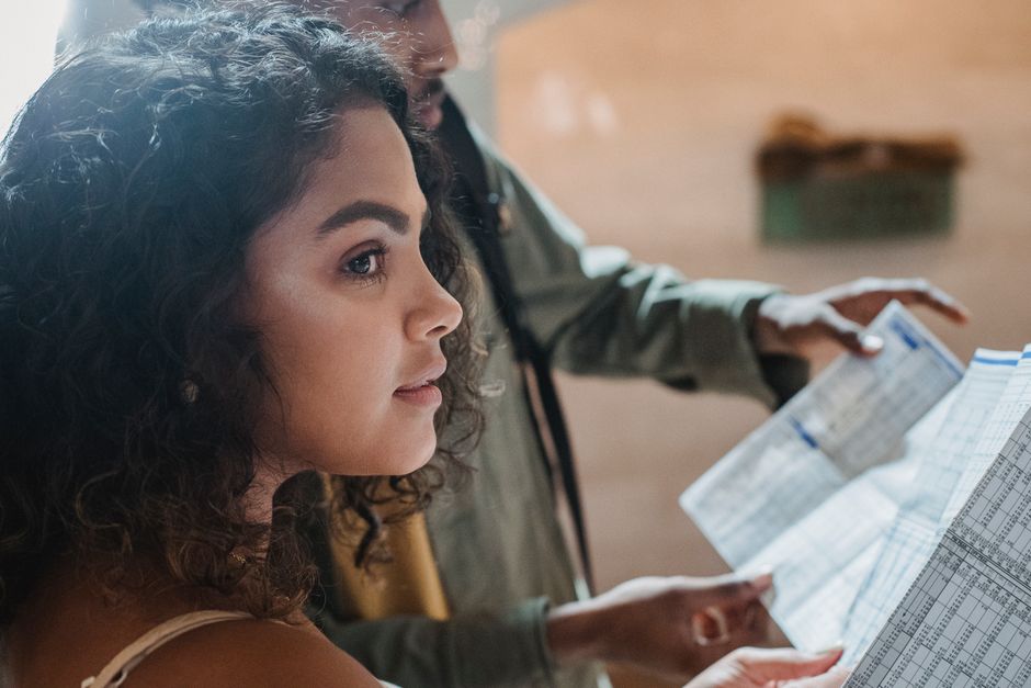 Side view of thoughtful Hispanic woman with dark curly hair standing with black friend reading long paper with tables