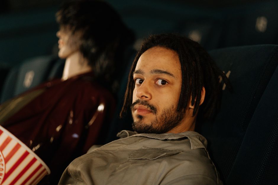 Man with dreadlocks and facial hair seated in a cinema, enjoying popcorn.