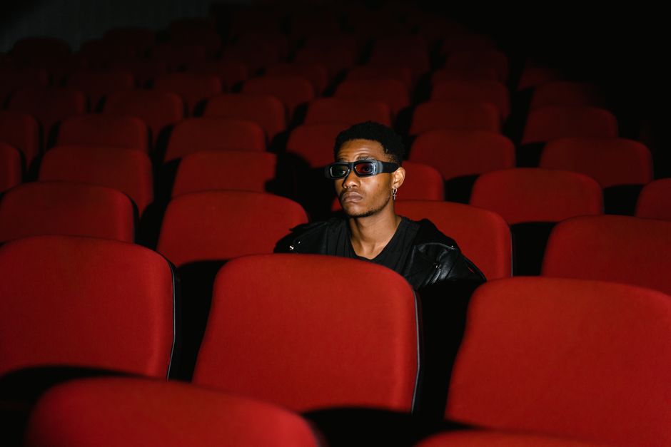 A man wearing 3D glasses sits alone in a movie theater with red seats, creating an isolated atmosphere.
