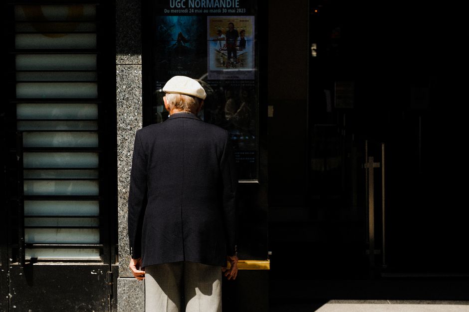Elderly man in classic attire viewing movie posters outside a cinema.
