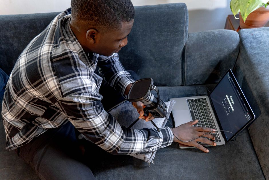 A young man holding a microphone and using a laptop on a sofa, indoors setting.