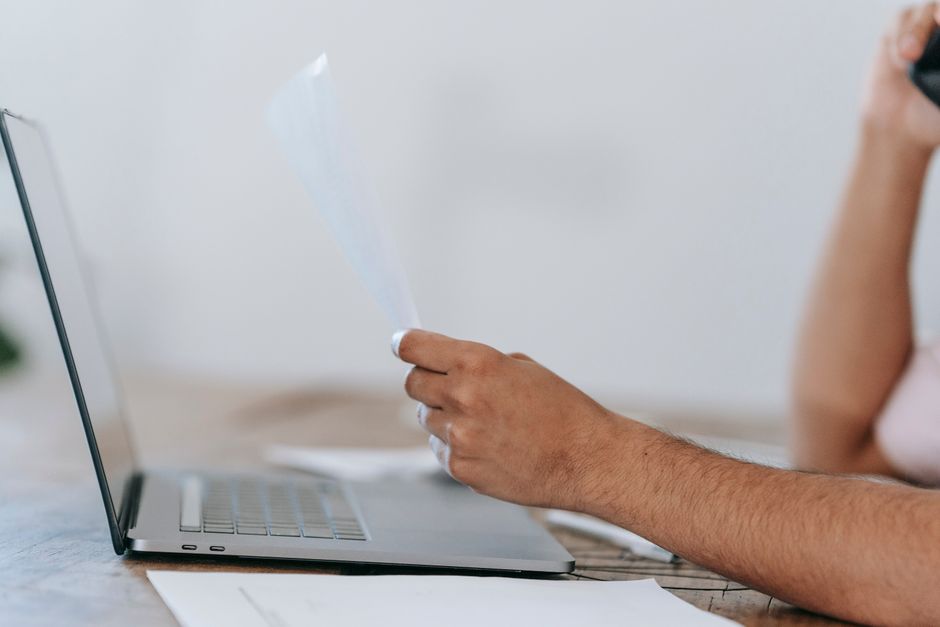 Crop anonymous male reading document and talking on mobile phone while sitting at desk with laptop
