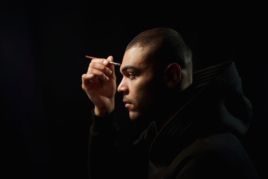A side view portrait of a man applying makeup in a dark studio setting.