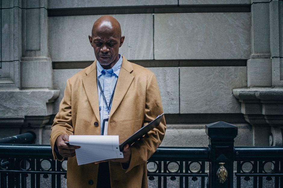 Serious black man in stylish clothes reading documents and standing on street near building in daytime