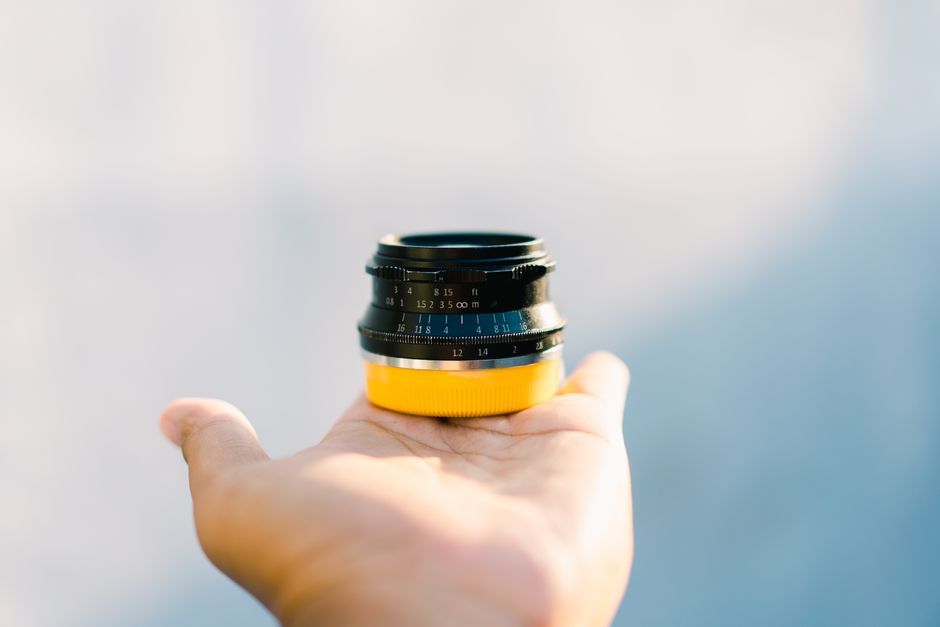 A hand holding a small camera lens, captured with soft focus outdoors.