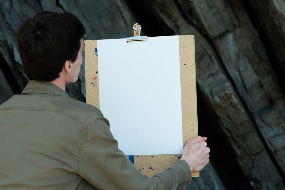 A man is setting up to paint on a blank canvas outdoors against a rocky backdrop.
