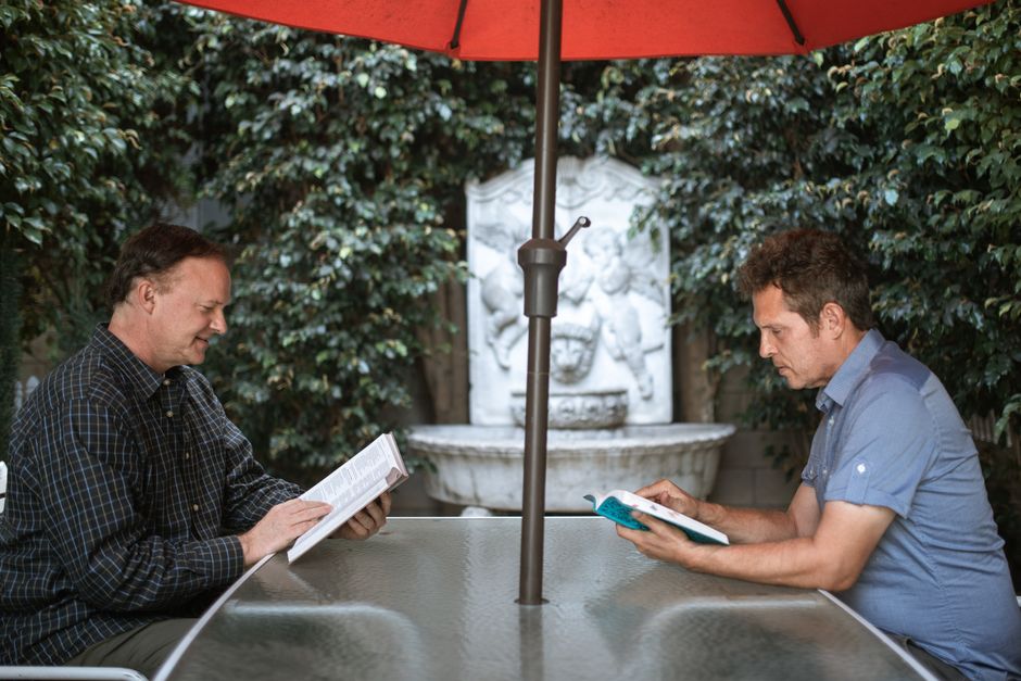 Two men enjoying a relaxed reading session at an outdoor table under a red umbrella.