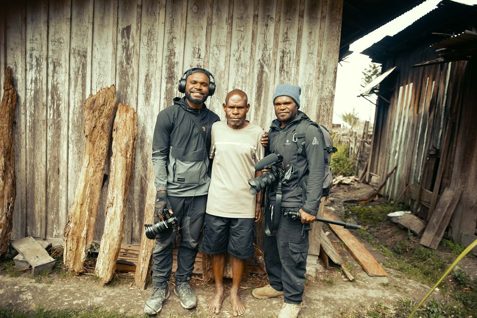 Three men, two with cameras, pose in front of a rustic wooden building outdoors.