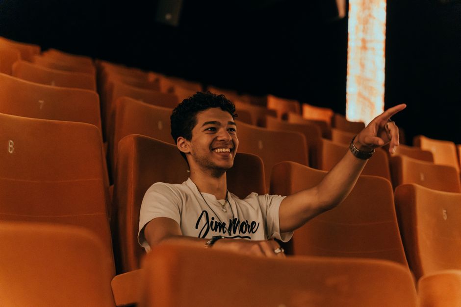 A young man with short hair smiles and points while seated in an empty cinema.