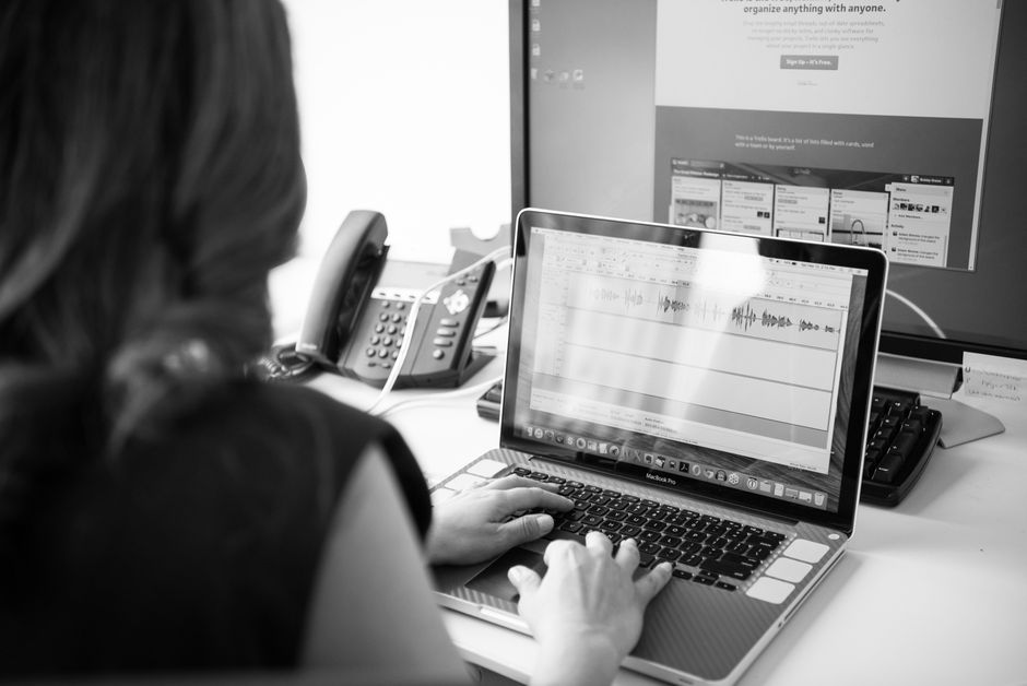 Black and white office scene with a woman working on a laptop in front of dual monitors.