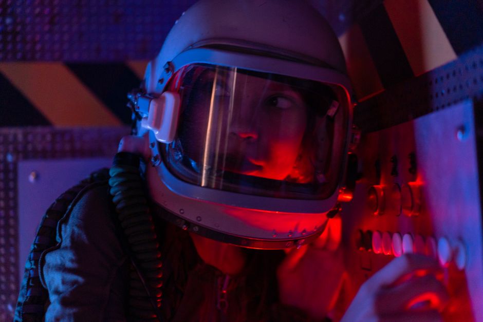 A woman astronaut wearing a spacesuit in a control room lit by red lights, looks focused.