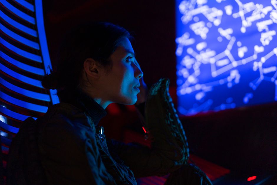 A woman in a space suit examines star maps in a futuristic, blue-lit environment, suggesting space exploration and technology.