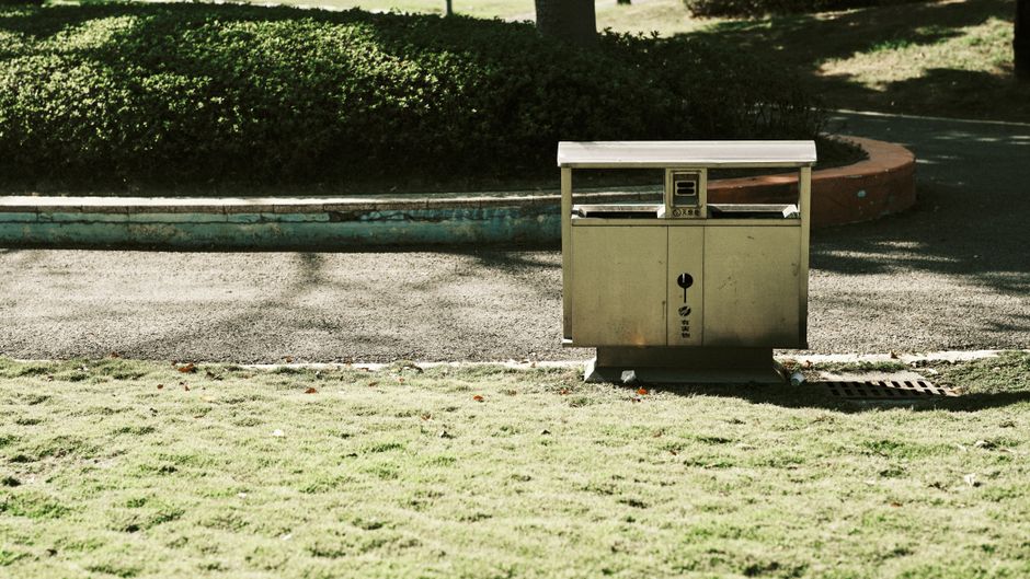 A solitary garbage bin in a quiet park surrounded by greenery and pavement.