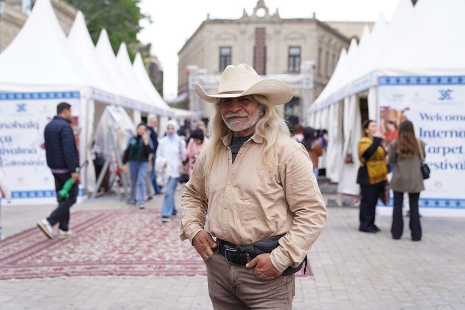 Man wearing a cowboy hat poses at an outdoor festival with numerous tents and people in the background.