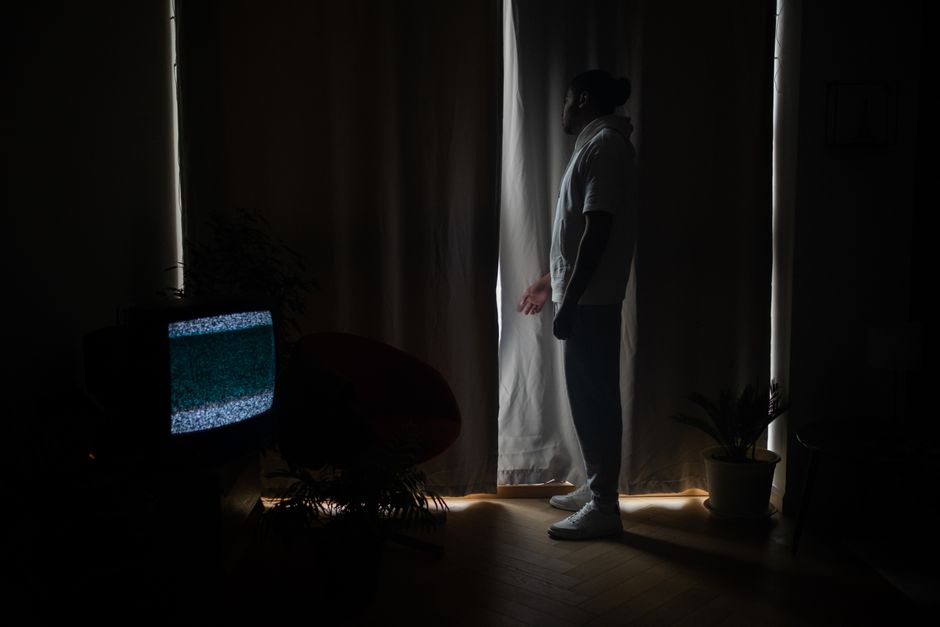 A man stands by curtains in a dimly lit room, television displaying static.