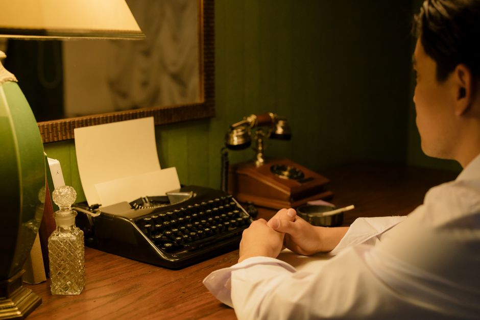A man in a vintage setting sits at a desk with a typewriter and lamp, reflecting a nostalgic workplace.