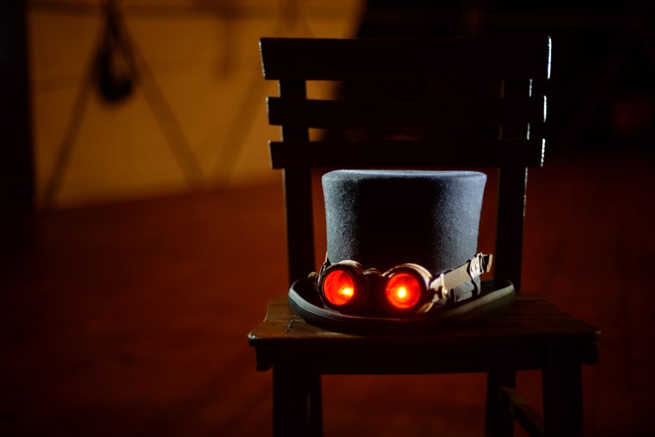 A steampunk-themed top hat with glowing goggles sits prominently on a wooden chair in dim lighting.