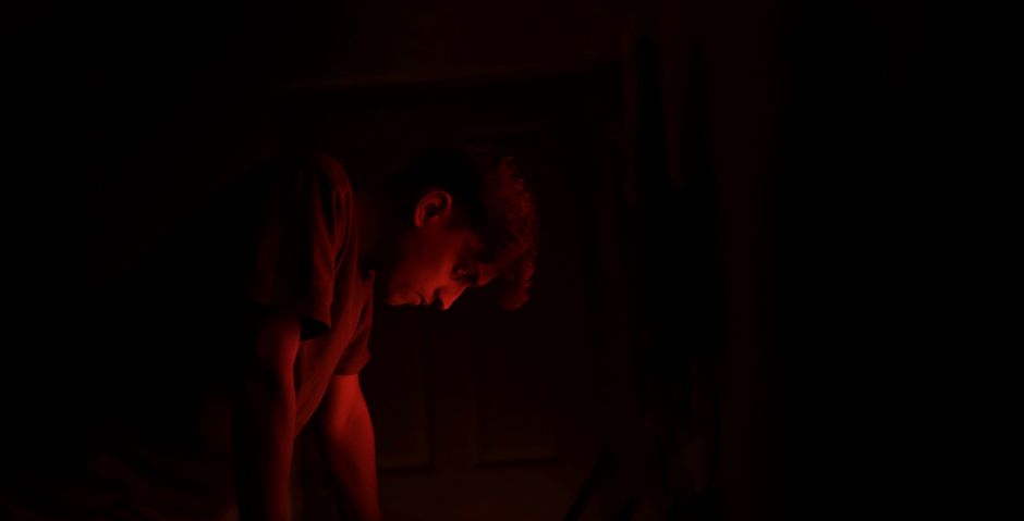 A man surrounded by deep shadows, illuminated by a warm red light, creating a mysterious atmosphere.