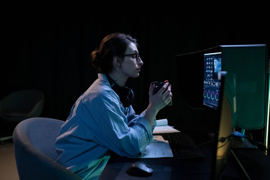 A woman in a dimly lit room working intently on a computer while holding a mug.