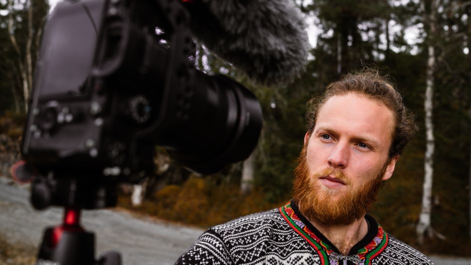 A bearded man records a video outdoors using a professional camera, showcasing selective focus and vlogging setup.