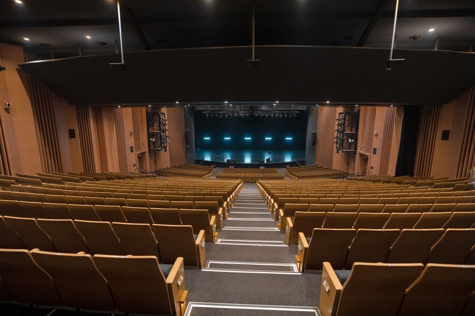 Wide view of an empty theater auditorium in Varanasi, India, featuring wooden chairs and a bright stage.