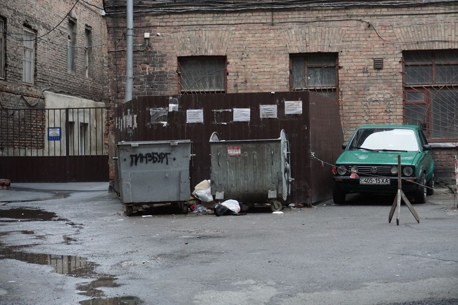 A gritty urban alley featuring garbage bins, scattered litter, and an old parked car.