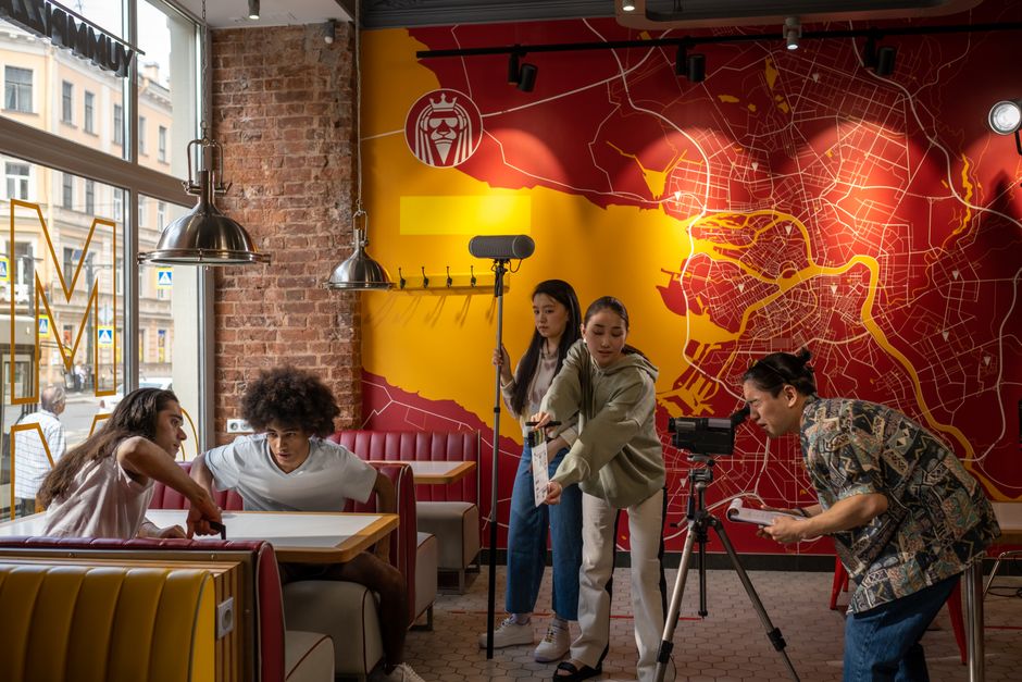 A diverse film crew operates in a trendy urban restaurant with a vibrant map mural.