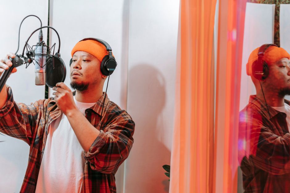 A young man in a recording studio adjusting a microphone while wearing headphones and a knit hat.
