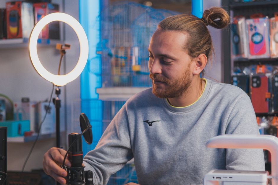 A man sitting in a studio setting adjusting a microphone with a ring light and equipment around.