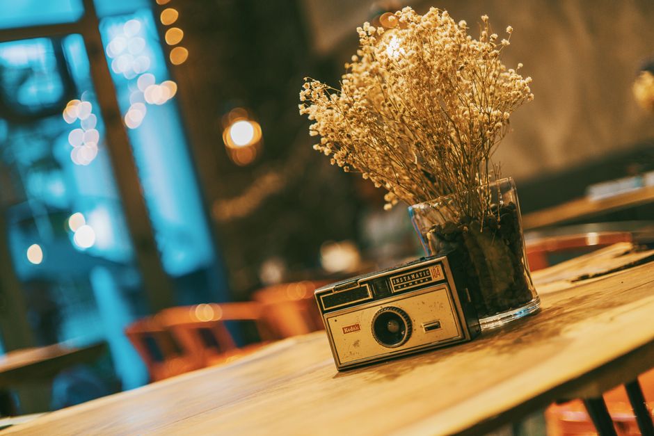 A vintage camera and dried flowers on a wooden table in a cozy indoor cafe setting.