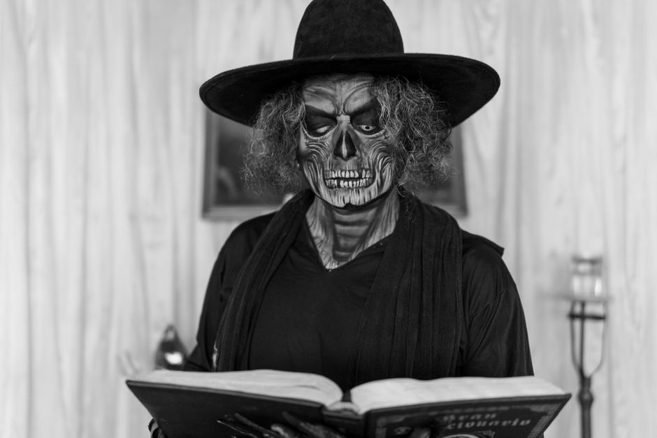 A person in costume with skull makeup reading an ancient book indoors, creating a mysterious atmosphere.