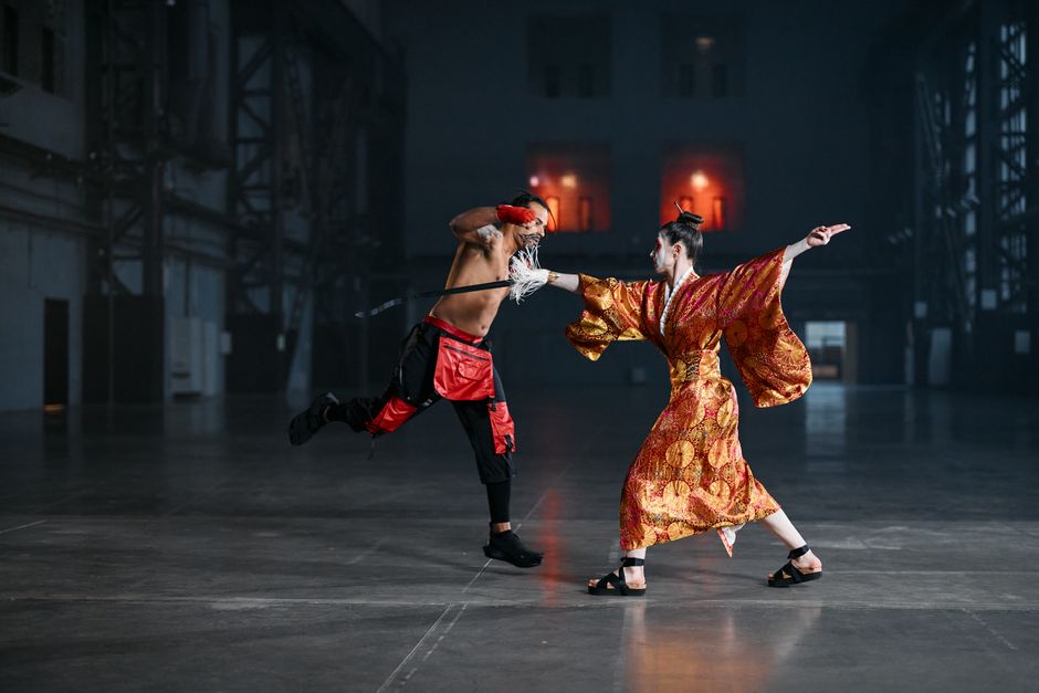 Two martial artists engage in a dramatic duel wearing traditional attire in a dimly lit setting.