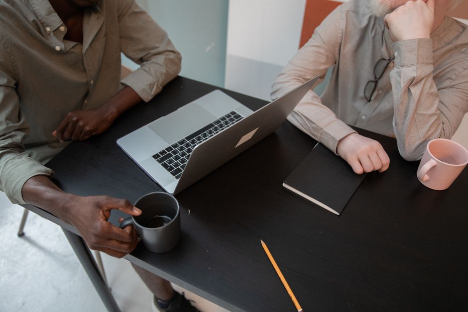 High angle of crop unrecognizable multiracial coworkers in casual clothes sitting at table with mugs of beverage and netbook while working together in office