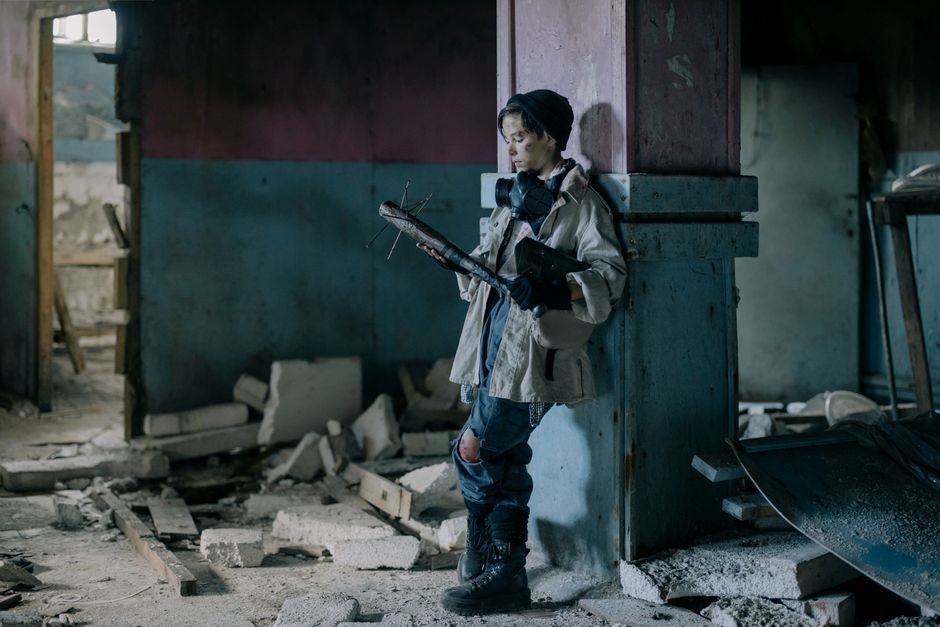 A young survivor in torn clothing observes a nail bat in a desolate, ruined setting.